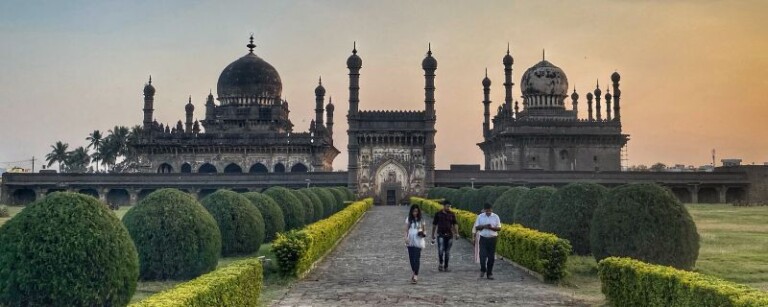 two indo persian style islamic buildings with garden in front with sky turned yellowish orange in bijapur