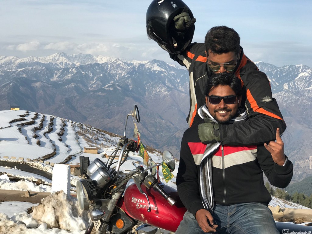 Indian male travel biker kissing onpillion rider's head sitting on royal enfield red bike with mountains and terraced land covered with snow behind 
