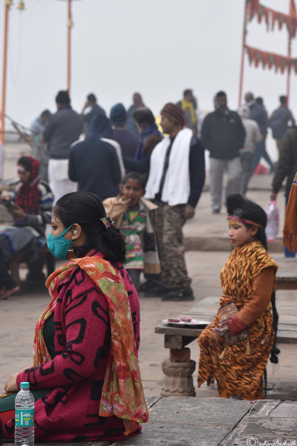 Indian woman travel blogger wearing mask and sitting by the Ganga Ghats next to a boy dressed as god Shiva
