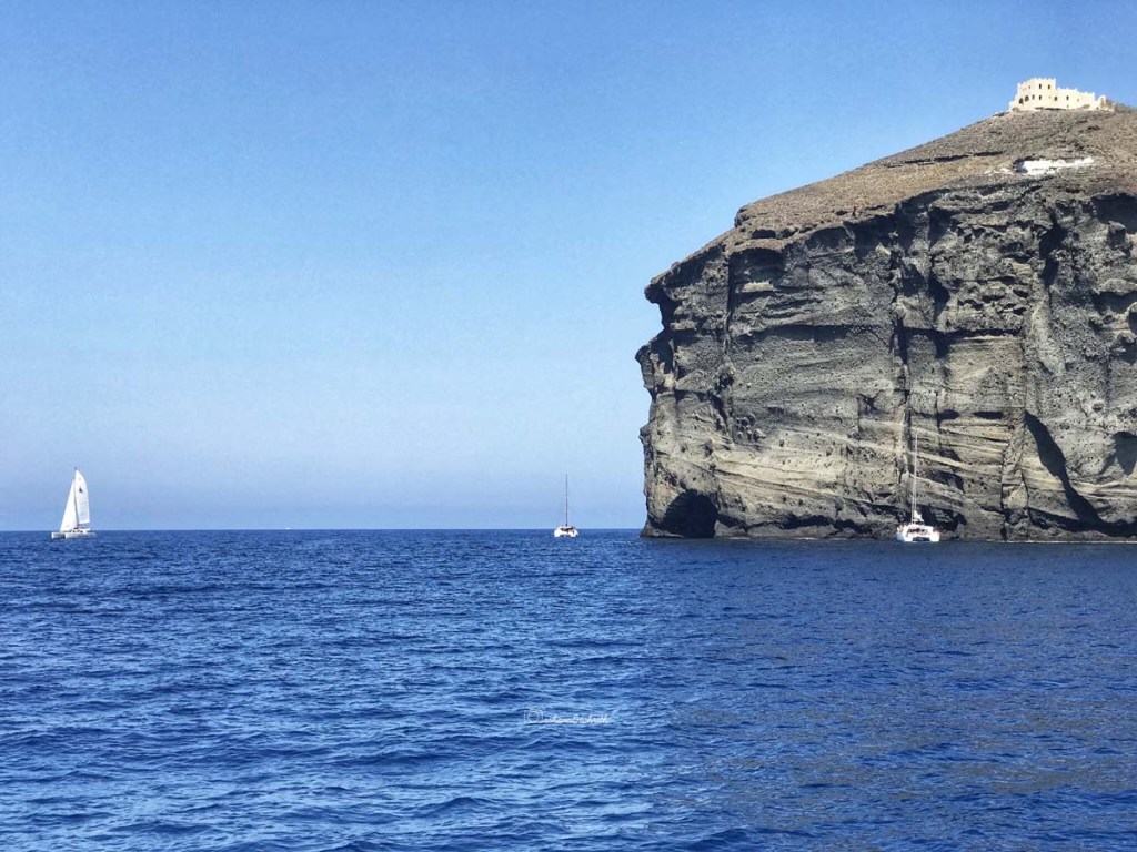 Dark blue water with yacths sailing on them beside a black mountain at Santorini