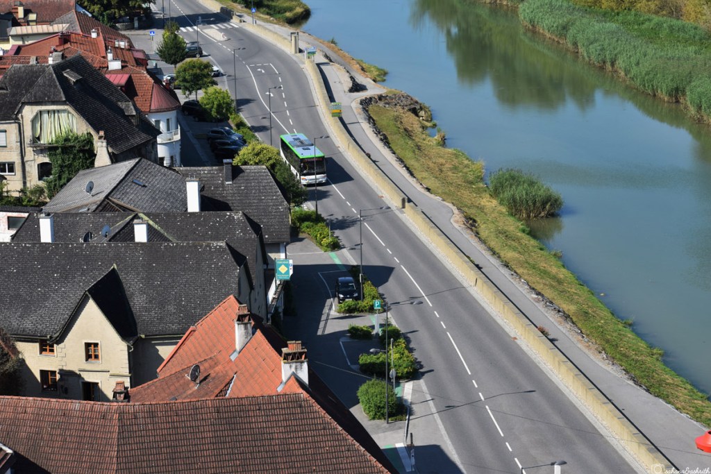 Aerial view of public bus running on asphalt road with melk river on one side and gabled roof houses with brown and black tiles on the other side in Wachau valley