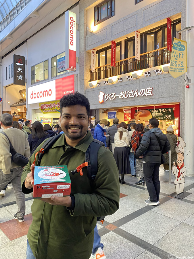 Indian man smiling and  holding box of packed Uncle rikuro jiggly cheesecake in the crowded street of osaka