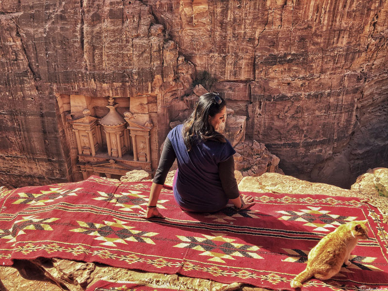 sahana sitting on a jordanian traditional carpet beside a cat at the cliff overlooking treasury of petra in Jordan