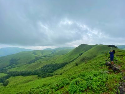 a girl standing with umbrella at the cliff edge of rolling green hills in chikmagalur