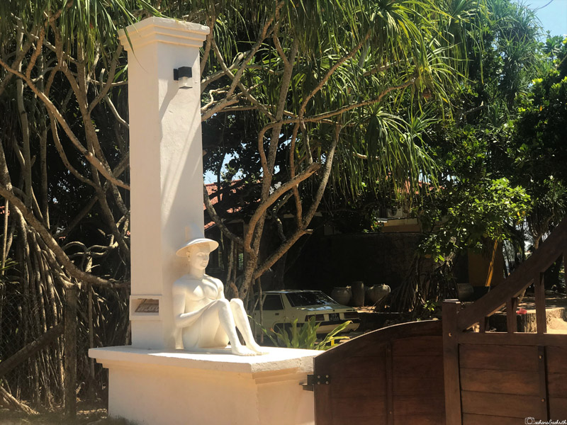 Statue of a man in a hat sitting and reading a book in front of a colonial bungalow