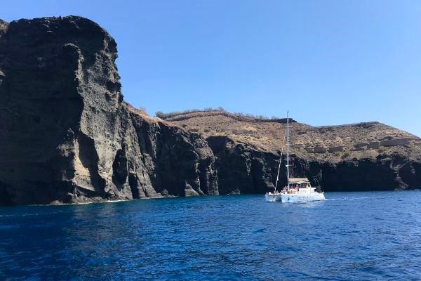 Catamaran sailing on vibrant Aegean waters alongside a brown volcanic peak in Santorini