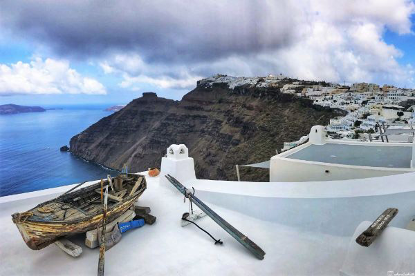 A classic boat beside a wall, with rows of white cube houses on one side and the Aegean Sea on the other, in Fira