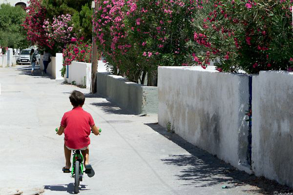 A young Greek boy riding his bicycle along a dirt road bordered by white houses and vibrant pink bougainvillea plants.