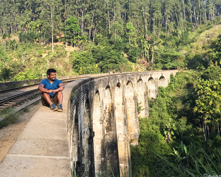 Indian man sitting on the parapet wall of train track above nine arch bridge in ella surrounded by tea estate