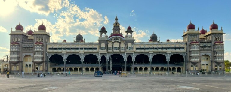 Panoramic view of Mysore Palace in daylight, highlighting its intricate architectural details and grandeur against a blue sky with scattered clouds