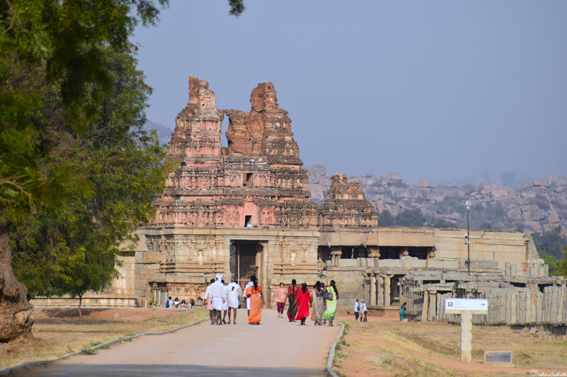 Visitors walking toward Hampi Vijaya Vittala Temple in gopuram ruin, split in half.
