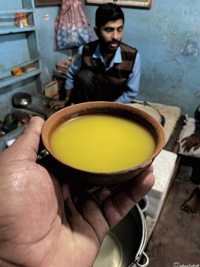 A hand holding a clay cup filled with a yellow liquid Thandai, with a man sitting in the background