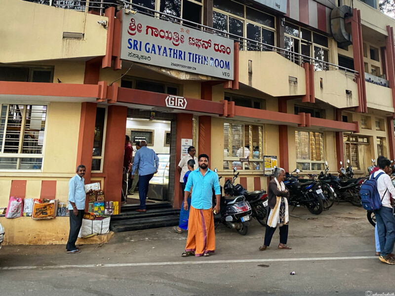 The entrance of Sri Gayathri Tiffin Room with people standing outside and a few scooters parked nearby