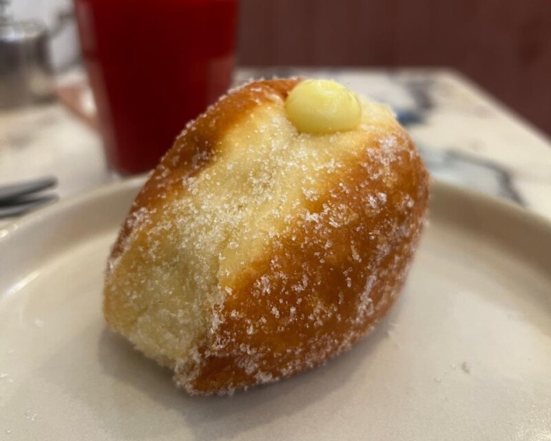 sugar-coated bomboloni doughnut filled with cream, placed on a white plate with a red cup in the background at SAPA bakery Mysore