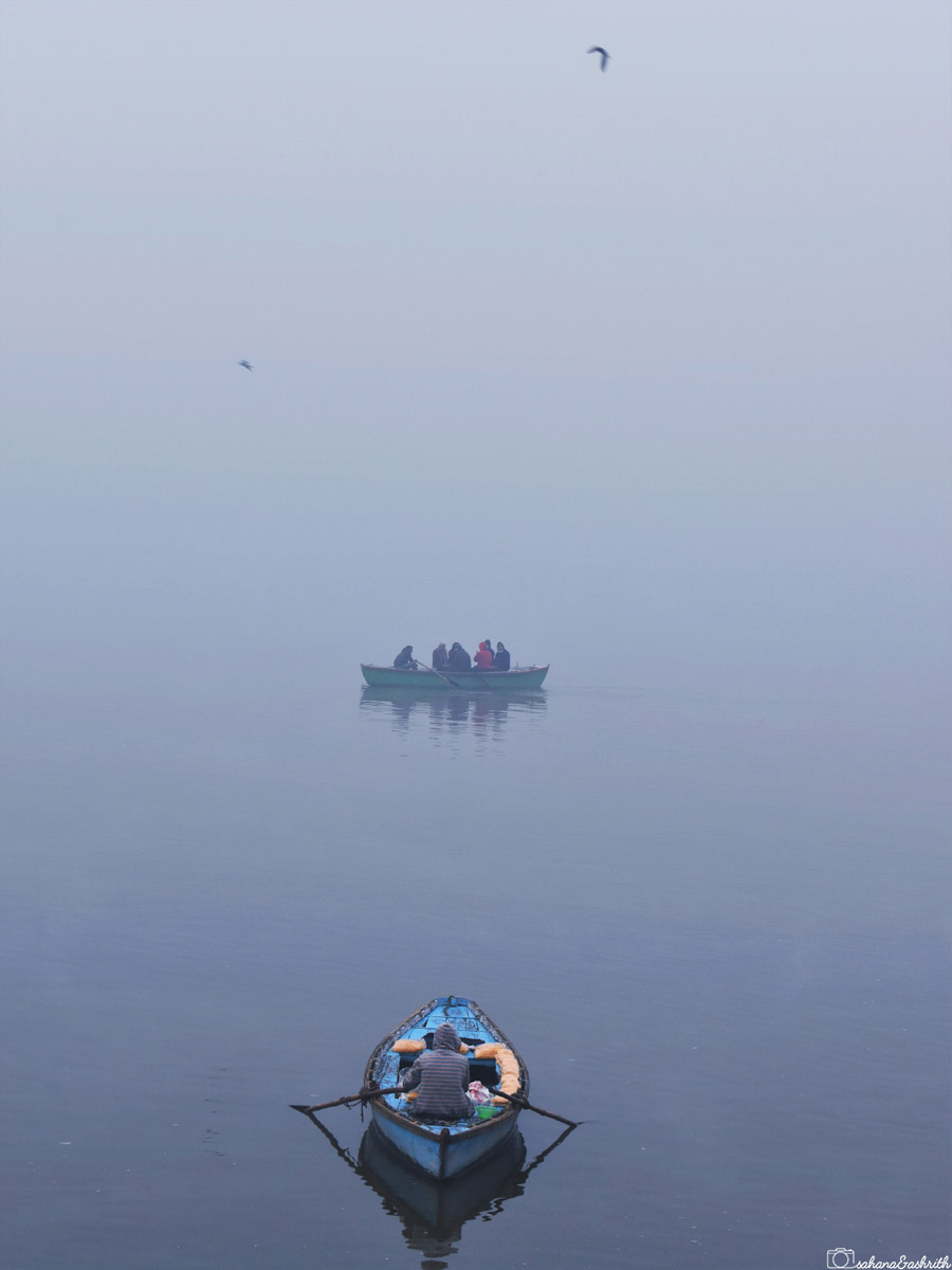 A misty lake scene with a solitary person rowing a blue boat in the foreground, and a group of people in another boat in the distance, with birds flying above