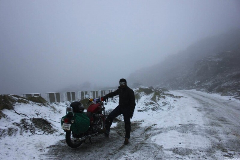 A motorcyclist, dressed in dark gear, stands next to their bike on a snowy and foggy mountain road