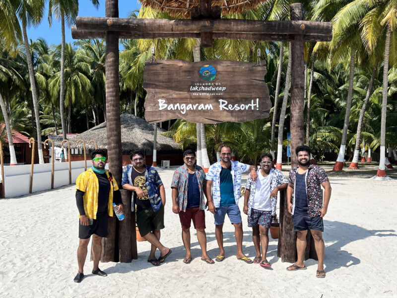 Six friends stand under a sign reading "Bangaram Resort" with palm trees and beach huts in the background.
