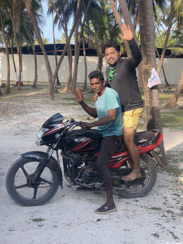 Indian tourist with lakshadweep local man on motorbike, waving and smiling, on a sandy path lined with palm trees