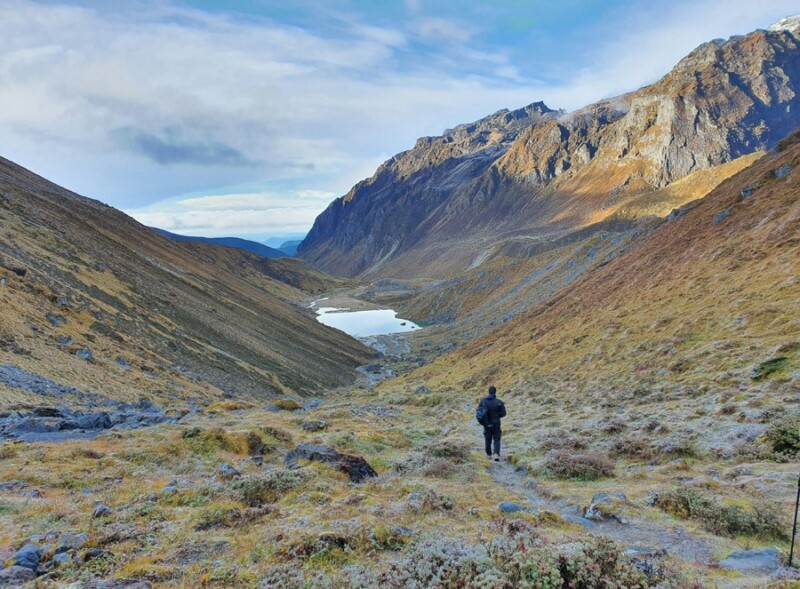 A person hikes through a scenic valley with towering mountains and a lake in the distance under a bright sky as part of his Goechela trek