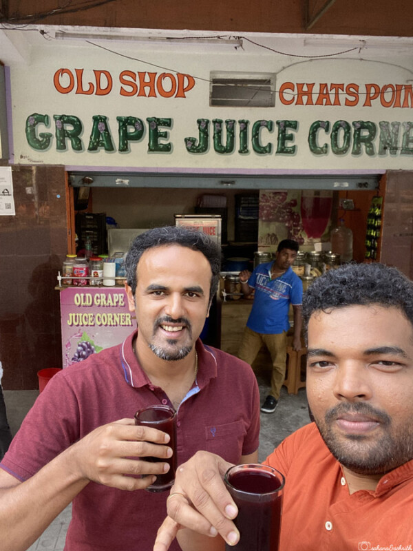 Two men smiling and holding glasses of grape juice in front of the Old Grape Juice Corner shop in Mysore