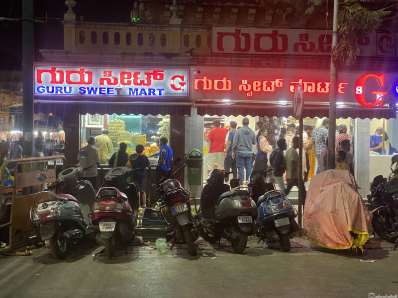 A night view of Guru Sweet Mart where mysore paks are stacked in shelfs with a crowd gathered outside and scooters parked in front.