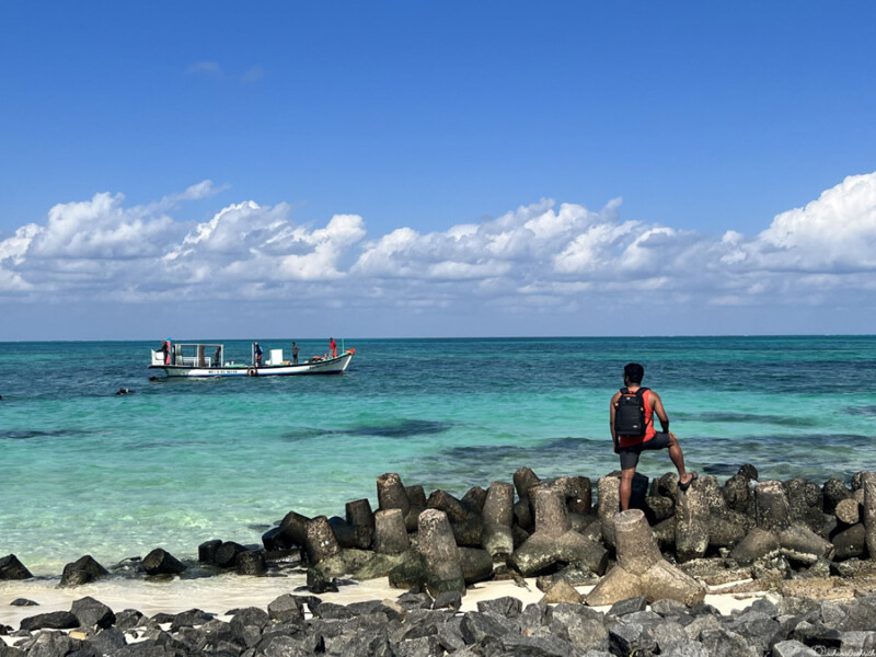 A man stands on large rocks by the shore, looking out at a boat on the turquoise waters under a bright blue sky.
