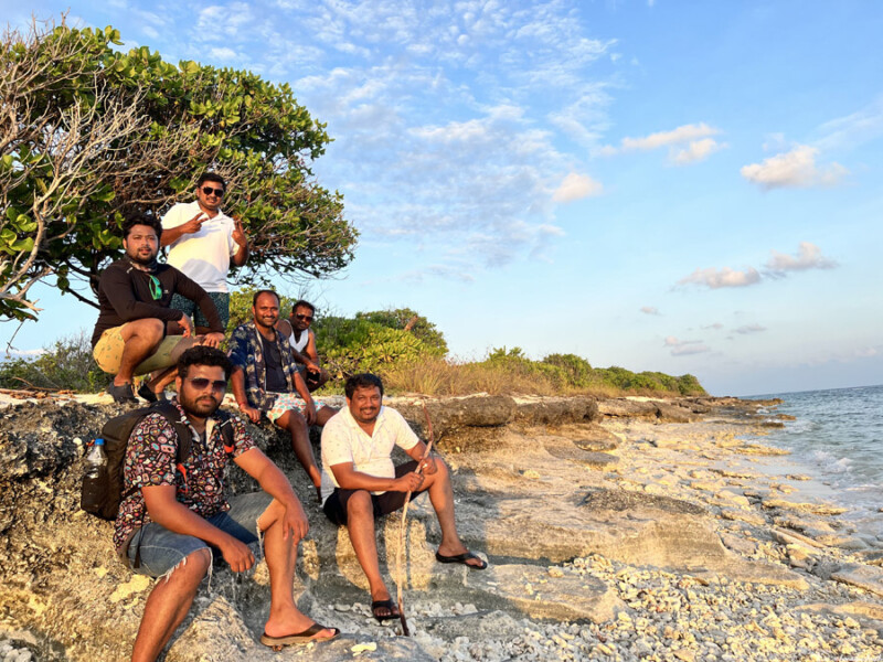 A group of friends sit on a rocky shore with the sea and a tree in the background