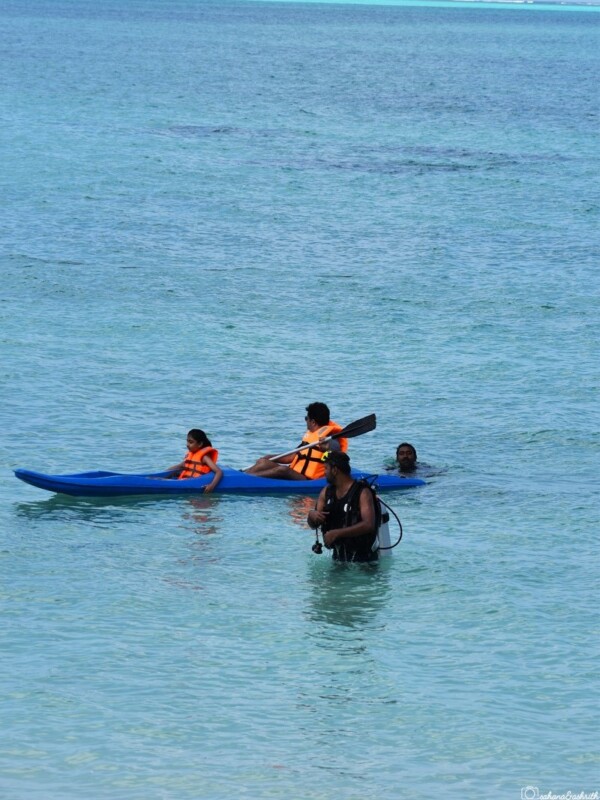 Two people, an adult and a child, both wearing life jackets, paddle a blue kayak together in the calm sea of Lakshadweep