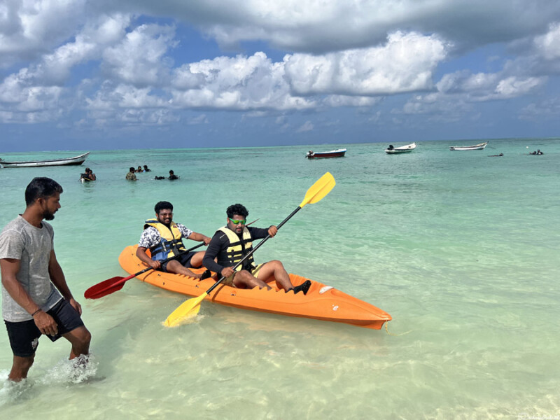 Two men paddle an orange kayak while a man walks along the shore in Lakshadweep