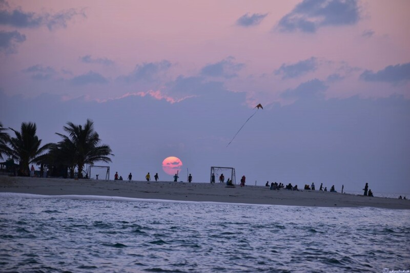 The sun sets over a beach, with people flying kites and playing in the distance.