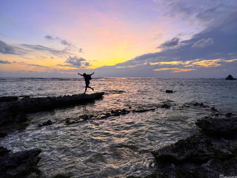 A person jumps from a rock into the sea at sunset.