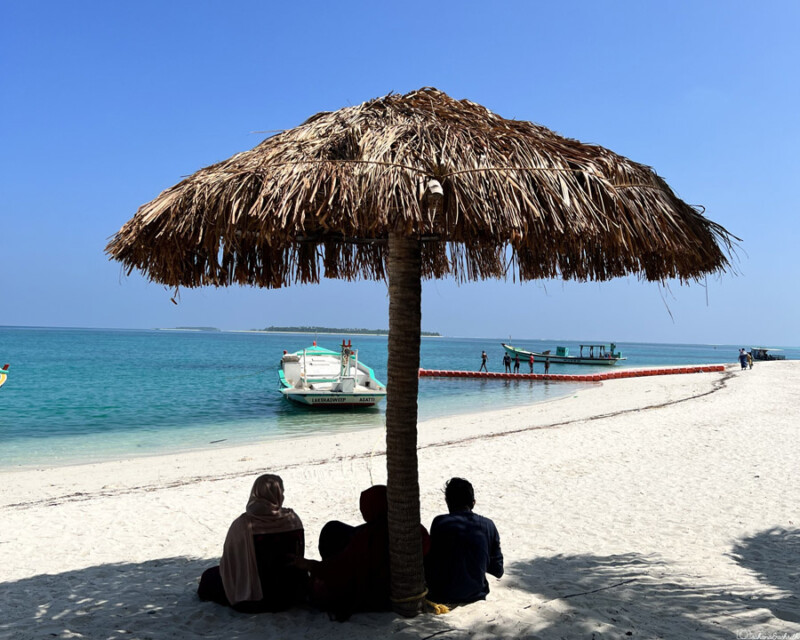 Three people, including two Muslim women wearing burqas, sit under a thatched beach umbrella, overlooking the turquoise waters and boats in Lakshadweep