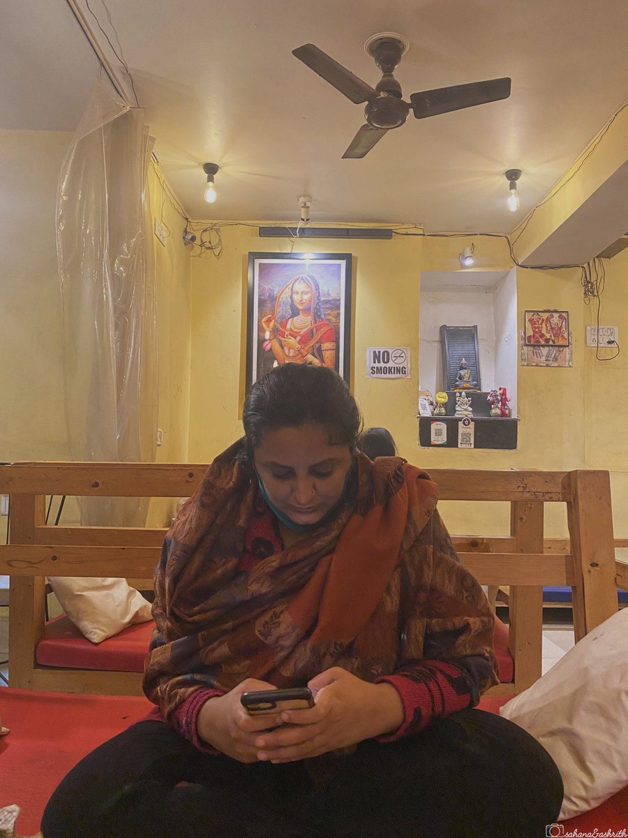 A woman sits on a wooden bench, focused on her phone, in a warmly lit room with yellow walls and a framed painting of an Indianised Mona Lisa wearing a ghagra choli, a ghoonghat over her head, and a nose ring.