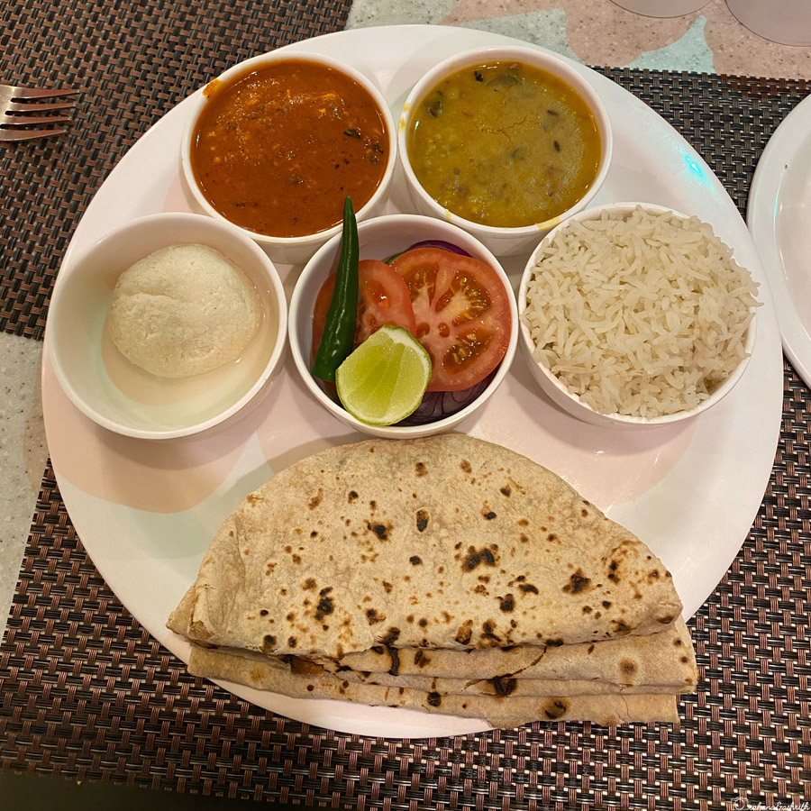 A traditional Indian meal served on a white plate, featuring bowls of dal, curry, rice, sliced vegetables, a green chilli, lime, and a sweet dessert, accompanied by chapati bread at Chulha Chauka