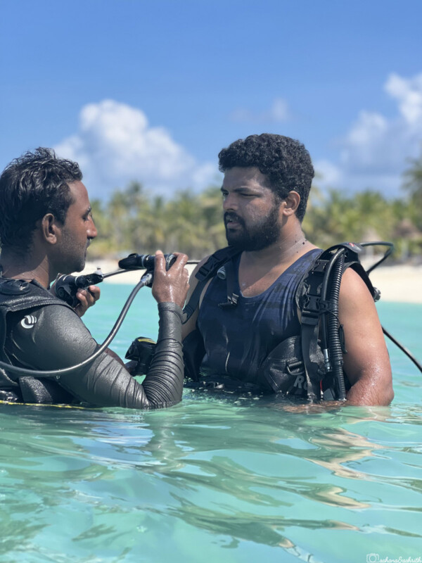 An instructor explains scuba diving equipment to ashrith standing in the shallow water