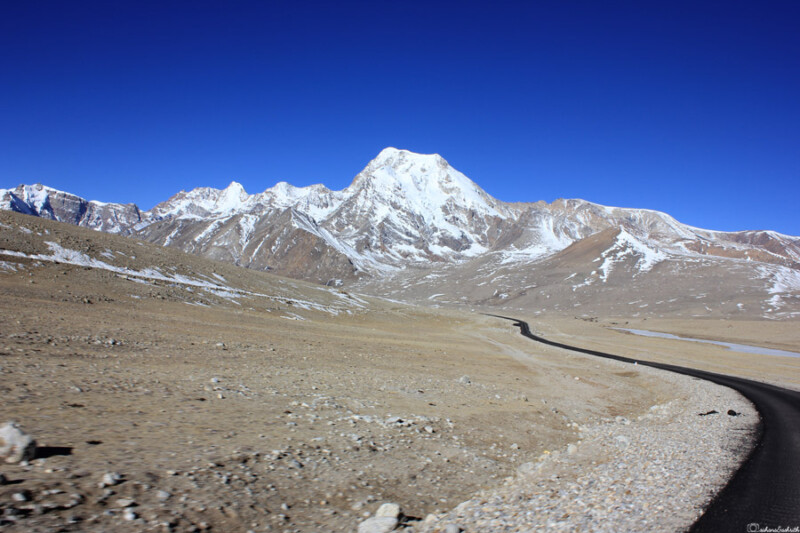 A paved road winds through a barren, mountainous landscape with snow-capped peaks under a clear blue sky.