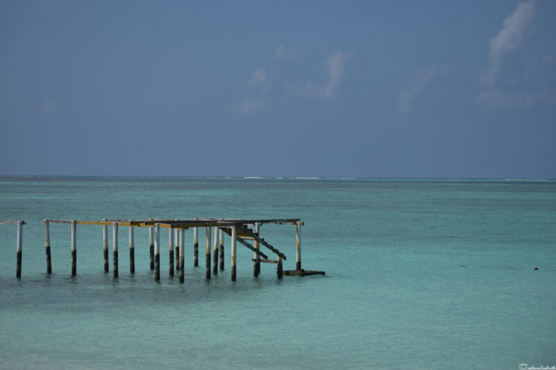 A dilapidated wooden pier extends into the calm, turquoise waters of the sea under a clear blue sky