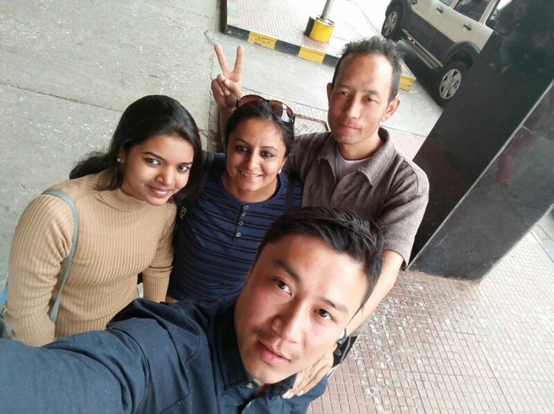 Two indian woman travellers taking a selfie with two sikkimese men with one person giving a peace sign, standing outdoors in a casual setting