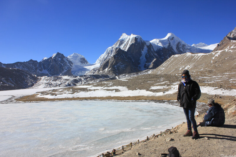 A person stands by a frozen lake with snow-covered mountains under a clear blue sky, dressed warmly against the cold.