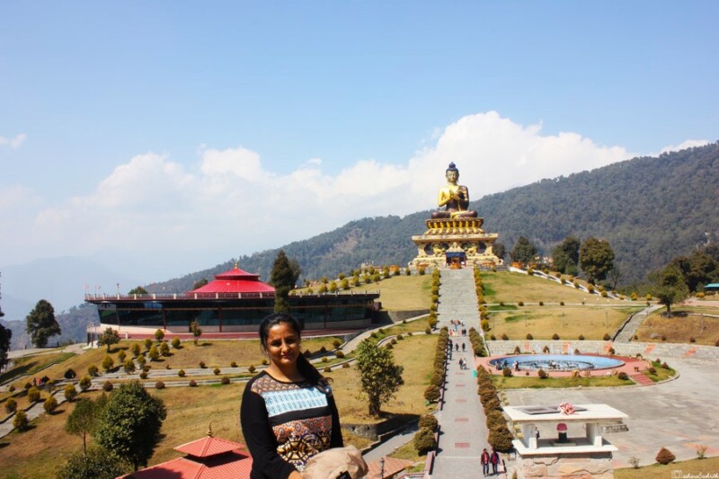 vacaywork author sahana posing in front of the towering Buddha statue at Buddha Park in Ravangla, South Sikkim, with a clear blue sky and surrounding mountains in the background
