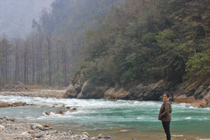 A woman wearing thick jacket, scarf stands on the rocky bank of a river with turquoise waters, surrounded by misty forested hills