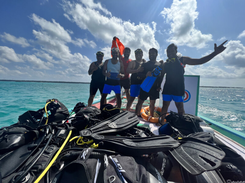 A group of six friends pose on a boat with their diving gear in the foreground.