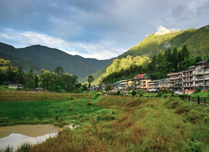 A serene remote village only accessible by private car with colourful buildings nestled in a lush green valley, surrounded by tall mountains under a partly cloudy sky.