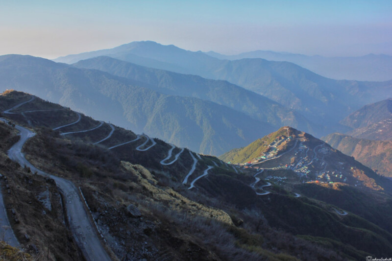 A winding road with multiple hairpin bends cutting through the rugged hills of Zuluk in East Sikkim, with layers of mountains fading into the distance under a clear sky