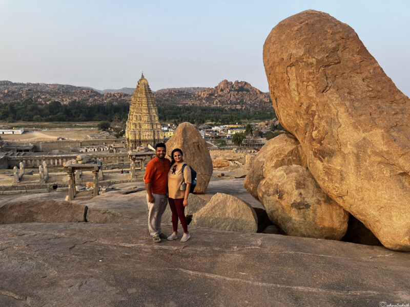 A couple standing on rocky terrain with the ancient Virupaksha Temple in the background.