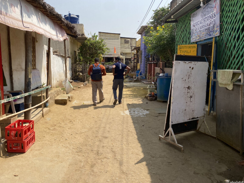 Two men walking down a narrow street in Hampi, India, with local shops and a signboard advertising South Indian Thali