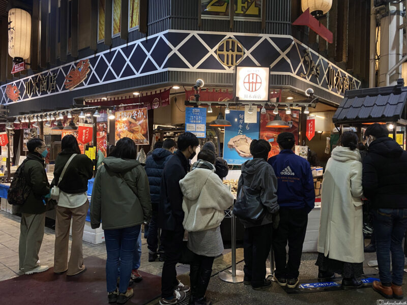 A group of people queueing at a seafood stall in a busy Japanese market,