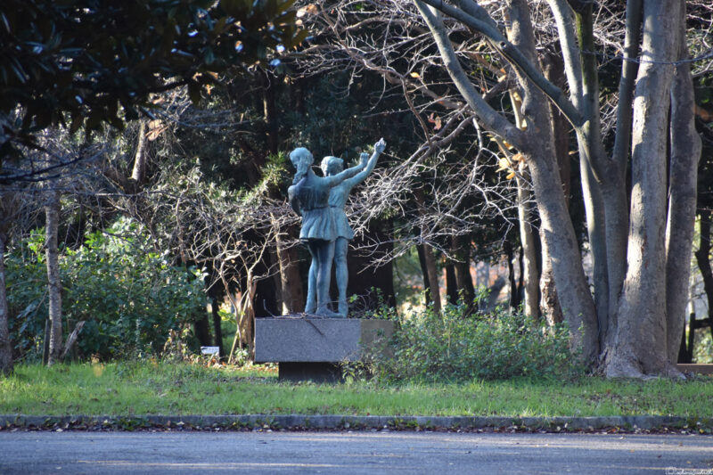 A statue of a couple pointing towards the trees in a park, surrounded by greenery and bare branches, capturing a serene moment.