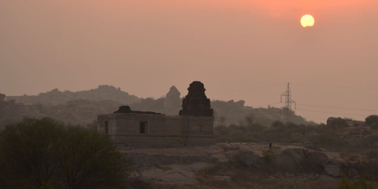 An ancient temple at sunrise with a glowing orange sun rising behind distant hills and a power line visible on the right.