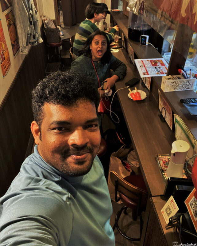  Ashrith taking a selfie in a restaurant; sahana behind him appears surprised, with diners in the background